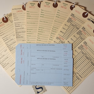 May include: A collection of vintage paper tags with handwritten text and numbers. The tags are used for tracking inventory, repairs, and alterations. The tags are beige and blue with black text.