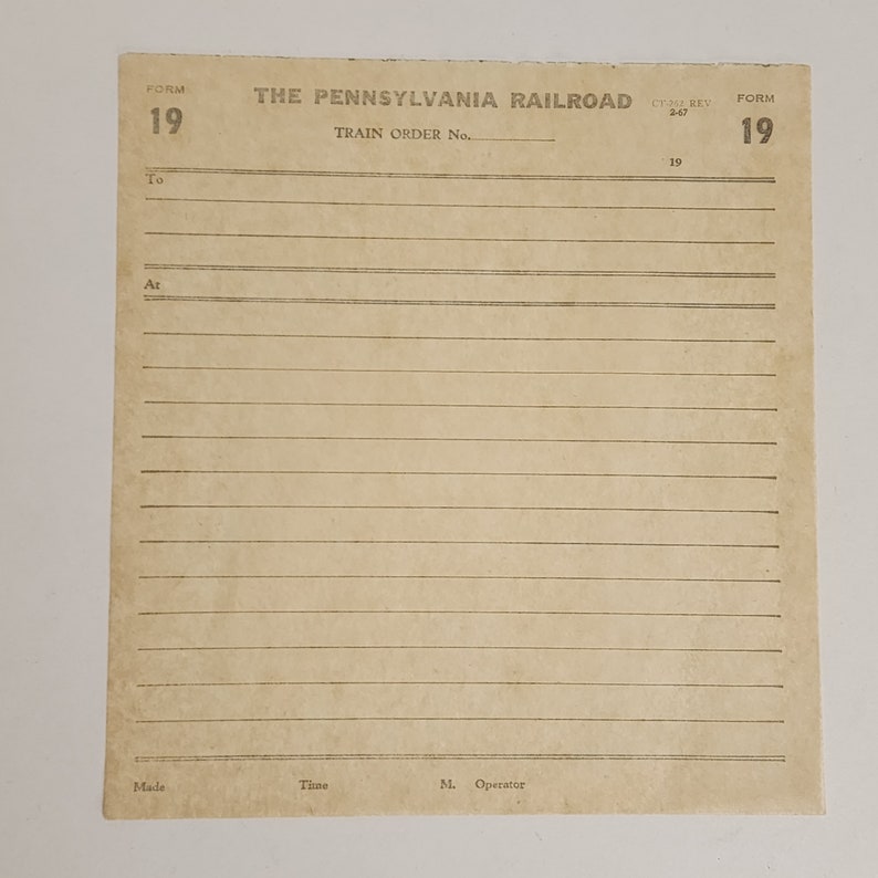 May include: A vintage train order form from The Pennsylvania Railroad. The tan paper has printed text and lines for writing. The form number is 19. The text includes "Train Order No.", "To", "At", "Made", "Time", and "M. Operator".