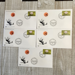 May include: Five white envelopes with stamps and postmarks. Each envelope features a black tree silhouette, an orange atom symbol, and text in French. Stamps are green and yellow with text. The envelopes are arranged on a light wood surface.