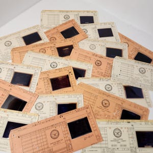 May include: A collection of vintage slides, featuring a mix of tan and off-white cardstock. Each slide has a rectangular window revealing a dark image. The cards are printed with text and a circular emblem.