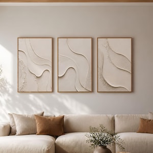 Puede incluir: Un conjunto de tres cuadros rectangulares con patrones abstractos de ondas texturizadas en color blanco roto dentro de marcos de madera clara, dispuestos horizontalmente sobre un gran sofá seccional beige con cojines marrones.