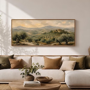 May include: A rectangular landscape painting in a wooden frame depicting a rolling hillside with olive trees, a dirt path, and distant mountains under a soft sky. The artwork is displayed on a plain wall above a white sofa with various pillows.