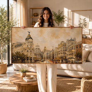 Puede incluir: Una impresión artística enmarcada que representa un paisaje urbano detallado con edificios, calles y luces. La obra de arte presenta una paleta de colores cálidos con tonos beige, marrón y dorado, creando una estética vintage. La impresión se sostiene en un salón bien iluminado.