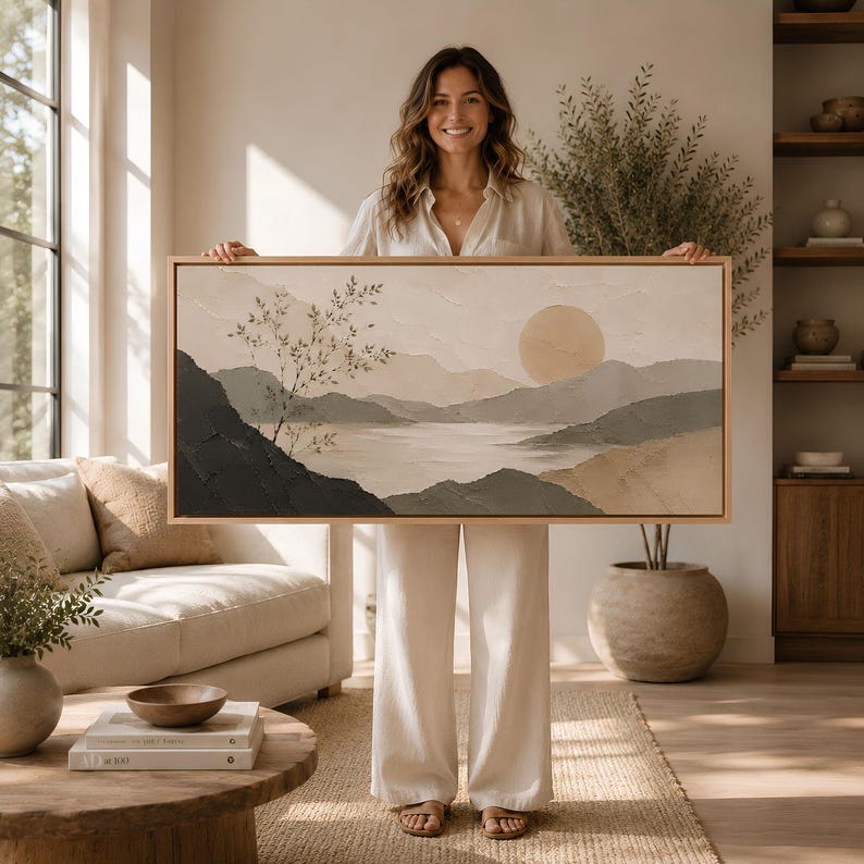May include: A rectangular landscape painting in a light wood frame featuring a textured scene of rolling hills, a calm body of water, and a large circular sun. The color palette consists of muted earth tones, including beige, cream, and dark gray.