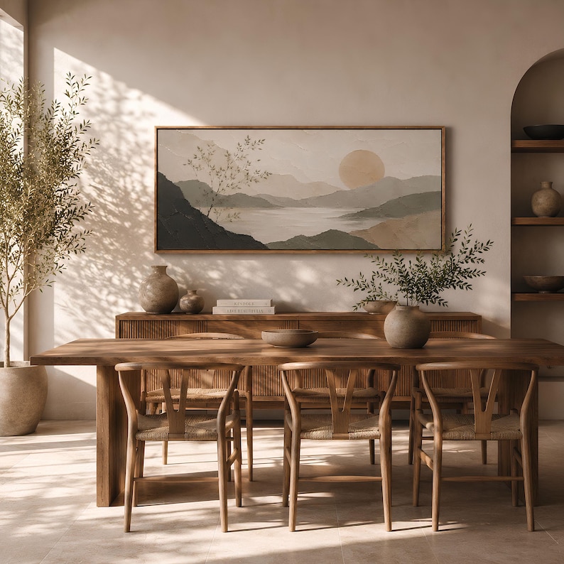 May include: A rectangular landscape painting in a wooden frame featuring a muted color palette of beige, green, and dark gray. The artwork depicts a serene mountain range, a calm body of water, and a large circular sun, creating a peaceful scene.