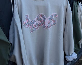 May include: A beige sweatshirt with the word "JESUS" appliquéd across the chest in a floral pattern. The long sleeves are cuffed, and the sweatshirt hangs on a black hanger against a wooden fence.