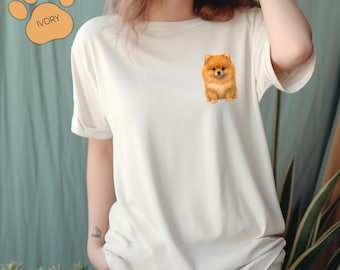 May include: Ivory-colored t-shirt featuring a small, detailed illustration of a Pomeranian dog on the chest. The shirt has short sleeves and a relaxed fit. A paw print graphic with the word "Ivory" is in the upper left corner.