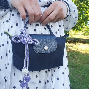 Puede incluir: Un bolso de mano azul marino con cierre de solapa y bot&oacute;n plateado. El bolso tiene un asa peque&ntilde;a y est&aacute; adornado con una borla decorativa morada y blanca. El fondo es un vestido blanco con lunares negros.