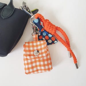 May include: A small orange and white gingham fabric pouch with a silver snap closure, attached to a silver metal lobster clasp. The accessory features a blue fabric bow with orange and white polka dots and a braided orange cord with silver tips.