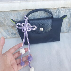 A black handbag with a leather flap and handle. A decorative purple and white knotted tassel with crocheted balls hangs from the handle. The bag has a silver button closure and lime green accents.