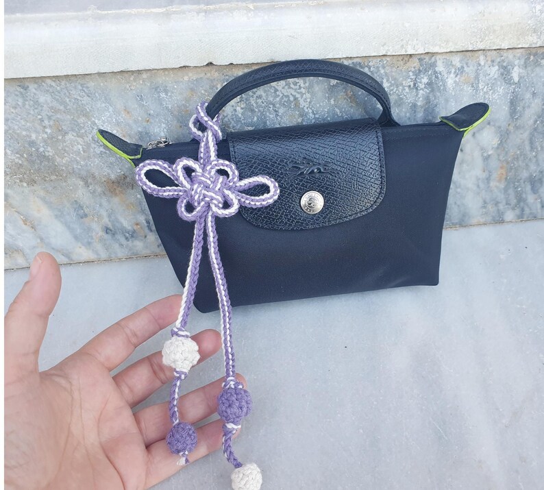 A black handbag with a leather flap and handle. A decorative purple and white knotted tassel with crocheted balls hangs from the handle. The bag has a silver button closure and lime green accents.
