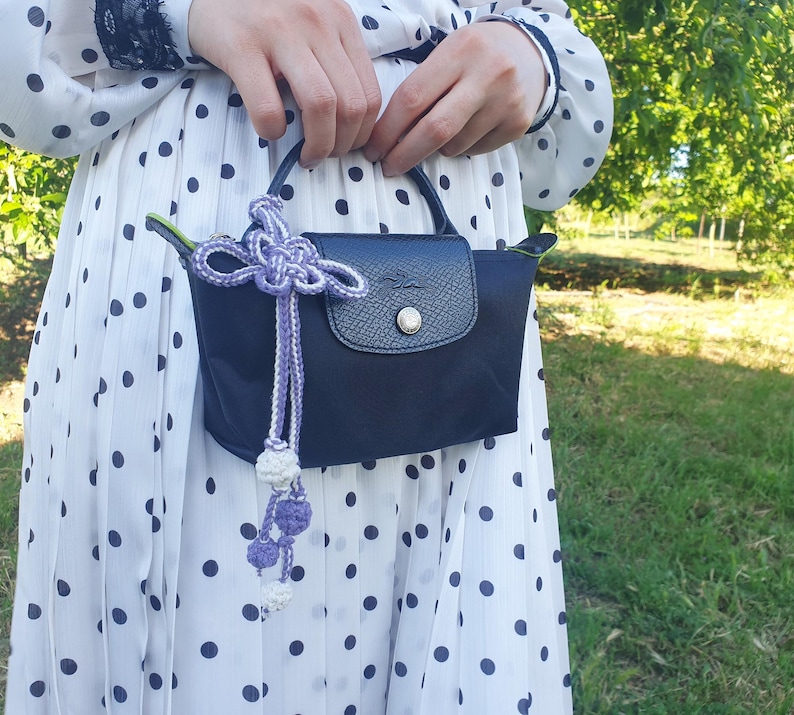 Puede incluir: Un bolso de mano azul marino con cierre de solapa y bot&oacute;n plateado. El bolso tiene un asa peque&ntilde;a y est&aacute; adornado con una borla decorativa morada y blanca. El fondo es un vestido blanco con lunares negros.