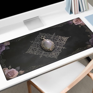 May include: A black desk mat with a floral and geometric design. The mat features a central ornate compass design and is adorned with roses in shades of pink and purple. A silver pendant with a gemstone is centered on the mat.