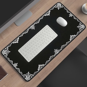 May include: A black rectangular desk mat with a white floral lace design border. A white keyboard and mouse sit on the mat. A brown notebook and a cup of coffee on a saucer are also visible on the wooden desk.