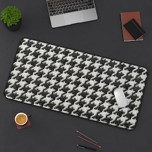 May include: A black and white houndstooth pattern desk mat with a black border. A white computer mouse sits on the mat. A laptop, a small potted plant, a notebook, and a cup of coffee are also on the dark grey surface.