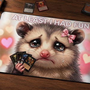 May include: A rectangular playmat featuring a digital illustration of a crying opossum with a pink bow on its ear, holding a hand of trading cards. The text AT LEAST I HAD FUN is printed in white at the top against a background of soft pink hearts.