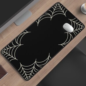 May include: A black rectangular mouse pad with a white spider web design along the edges. A white computer mouse sits on the pad. The pad is on a wooden desk, with a cup of coffee and a monitor in the background.