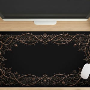 May include: Black rectangular mouse mat with a decorative bronze border featuring a thorny vine design. A white computer mouse rests on the mat, which sits on a wooden desk. A computer monitor and a small cactus are also visible.