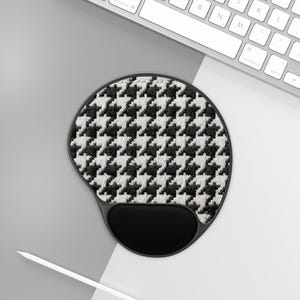 May include: A black and white houndstooth pattern ergonomic mouse pad with a black wrist rest. The mouse pad is round with a curved bottom and is positioned near a white keyboard and a white pencil.