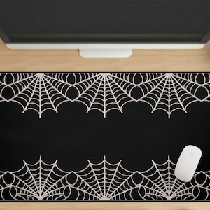 May include: Black rectangular mousepad featuring a white spiderweb design along the top and bottom edges. A white computer mouse rests on the right side of the pad. The mousepad is on a wooden desk.