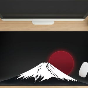 Puede incluir: Alfombrilla de escritorio rectangular negra con una ilustración blanca de una montaña y un sol rojo. Un ratón de ordenador blanco está a la derecha. La alfombrilla está sobre un escritorio de madera. Mide unos 80 cm de largo.
