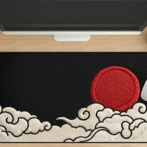 Puede incluir: Alfombrilla de ratón rectangular negra con un diseño bordado que presenta un sol rojo y nubes blancas. Un ratón de ordenador blanco está en el lado derecho de la alfombrilla. La alfombrilla está sobre un escritorio de madera.