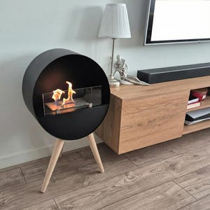 Op de afbeelding: Een moderne, zwarte, cirkelvormige open haard met een glazen voorkant, met een warme vlam. De open haard wordt ondersteund door drie houten poten. Het ontwerp is minimalistisch en eigentijds, geschikt voor binnenshuis. Een stijlvol woondecoratie-item.