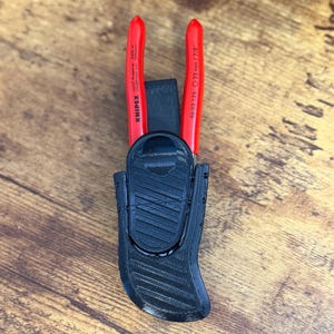 Può includere: Una pinza Knipex con manici rossi e la scritta 23 mm / 7/8" sul manico, fissata all'interno di una fondina nera stampata in 3D. La fondina presenta una superficie testurizzata con scanalature diagonali e un meccanismo a clip circolare frontale.
