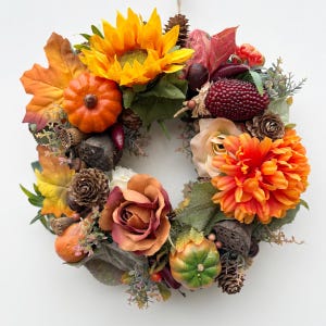 May include: A circular autumn wreath featuring a bright yellow sunflower, an orange dahlia, and a deep rose-colored fabric flower. The arrangement includes small orange and green pumpkins, brown pinecones, dried seed pods, and maple leaves.