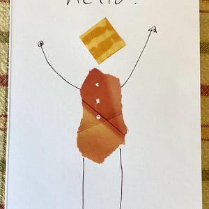 May include: A white greeting card with the word "hello!" in black script. The card features a whimsical figure made of torn paper in shades of orange and brown, with arms and legs drawn in black. A square of yellow paper serves as the head.