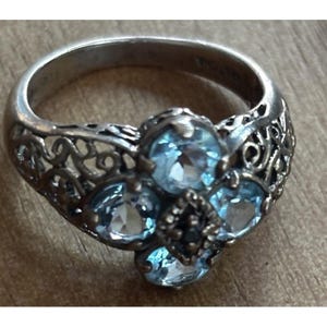 May include: Silver-toned ring featuring a cluster of light blue gemstones. The ring has an ornate filigree design on the sides and a central arrangement of four larger and one smaller gemstone. The gemstones are round-cut.