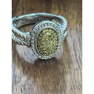May include: A silver ring with an oval-shaped center stone. The center stone is a mosaic of small, light green gems. The ring has a twisted rope design on the band and a halo of small, clear gems around the center stone.