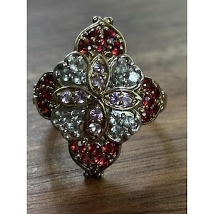 Puede incluir: Un anillo dorado con un diseño floral. El anillo presenta una forma de flor central con piedras preciosas rosas claras y transparentes, rodeadas de piedras preciosas rojas. El anillo está sobre una superficie de madera.