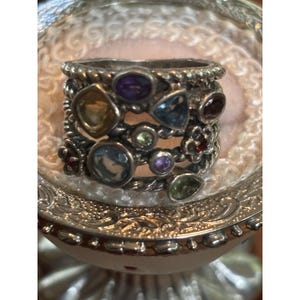 Può includere: Un ampio anello in argento con un design a fascia in corda intrecciata, decorato con molteplici pietre preziose multicolori di varie forme, tra cui pietre rotonde, ovali e a goccia nelle tonalità viola, blu, giallo, verde e rosso scuro.