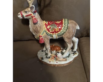 May include: A ceramic reindeer figurine with a decorative saddle and red tassels. The reindeer has large antlers and is standing on a base with small woodland creatures. The figurine is painted in shades of brown, red, green, and gold.