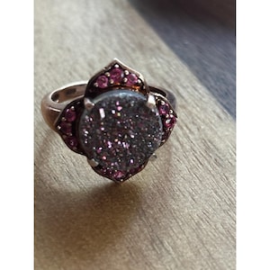 Puede incluir: Un anillo de oro rosa con una piedra central grande, redonda, brillante y gris oscuro. El anillo está rodeado de piedras preciosas rosas más pequeñas y redondas y tiene un diseño floral decorativo.
