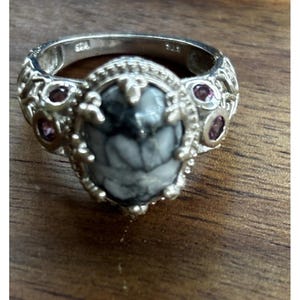 Puede incluir: Un anillo de plata con una piedra ovalada gris y blanca en el centro, flanqueada por piedras moradas más pequeñas y redondas. El anillo tiene detalles ornamentados en la banda y alrededor de la piedra central, de estilo vintage o antiguo.