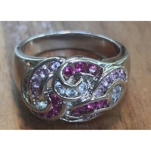 May include: A silver-toned ring featuring a swirling, openwork design set with multiple small, round-cut gemstones in shades of deep magenta, light pink, and clear white. The metal band is smooth and polished, showcasing a continuous, intricate pattern.