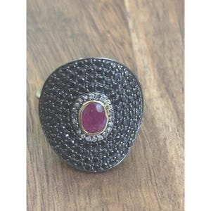 May include: A ring featuring a large, circular, dark metal face densely encrusted with numerous small black stones. At the center, an oval-shaped, deep pinkish-red gemstone is surrounded by a thin border of tiny, clear, sparkling white stones.