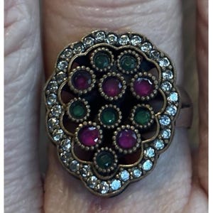 Puede incluir: Anillo ornamentado con un centro oscuro, con piedras de color rubí y esmeralda alternadas. El anillo está rodeado por un borde de cristales claros y brillantes, engastados en un metal dorado.