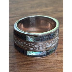 May include: A wide band ring featuring a central silver-toned metal band with an embossed swirling scroll pattern, flanked by two horizontal rows of iridescent abalone shell inlay with dark green, blue, and purple tones set in a polished metal frame.