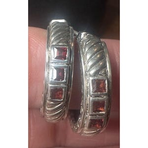 May include: A pair of silver hoop earrings. Each earring features three square, red gemstones set within a textured, braided design. The earrings are shown close-up, highlighting the details of the metalwork and the gemstones.