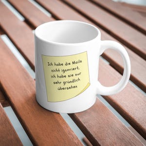May include: A white ceramic mug featuring a yellow sticky note graphic on the front. The note contains the German text: Ich habe die Mails nicht ignoriert, ich habe sie nur sehr gründlich übersehen. The mug has a standard handle and a smooth finish.
