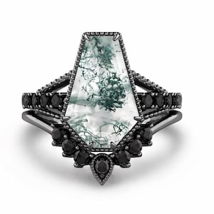 May include: A ring set featuring a central coffin-shaped moss agate stone with green dendritic inclusions, set in a black metal band. The setting is accented with small, round, faceted black stones along the band and a curved, beaded halo.