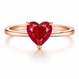 May include: Rose gold ring featuring a heart-shaped, faceted red gemstone. The gemstone is the focal point, held in place by four small prongs. The band is a smooth, polished rose gold.