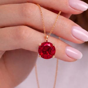 Op de afbeelding: Een goudkleurige ketting met een grote, ronde, felrode edelsteen hanger. De hanger wordt op zijn plaats gehouden door vier kleine gouden pootjes. De ketting is delicaat en lijkt ook goud te zijn. De ketting wordt tegen een zachte achtergrond getoond.