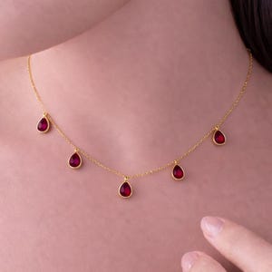 Könnte beinhalten: Eine zarte goldene Halskette mit sechs tropfenförmigen, tiefroten Edelsteinen. Die Edelsteine sind gleichmäßig an der feinen Goldkette angeordnet. Die Halskette wird vor einem sanften rosa Hintergrund präsentiert.