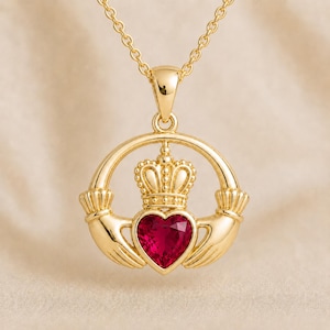May include: A gold-colored Claddagh pendant necklace. The pendant features a heart-shaped red gemstone, a crown, and two hands. The necklace hangs from a delicate gold chain. The Claddagh design is a symbol of love, loyalty, and friendship.