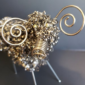 Può includere: Una scultura in metallo argentato di una pecora con un corpo di lana di filo metallico riccio e gambe di filo metallico. La pecora ha due grandi corna di filo metallico ricci e due piccole perline rotonde d'argento per gli occhi.