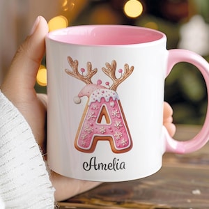 May include: White ceramic mug with a pink interior and handle. The mug features a pink and brown gingerbread-style letter "A" decorated with snowflakes and antlers. The name "Amelia" is written below the letter.
